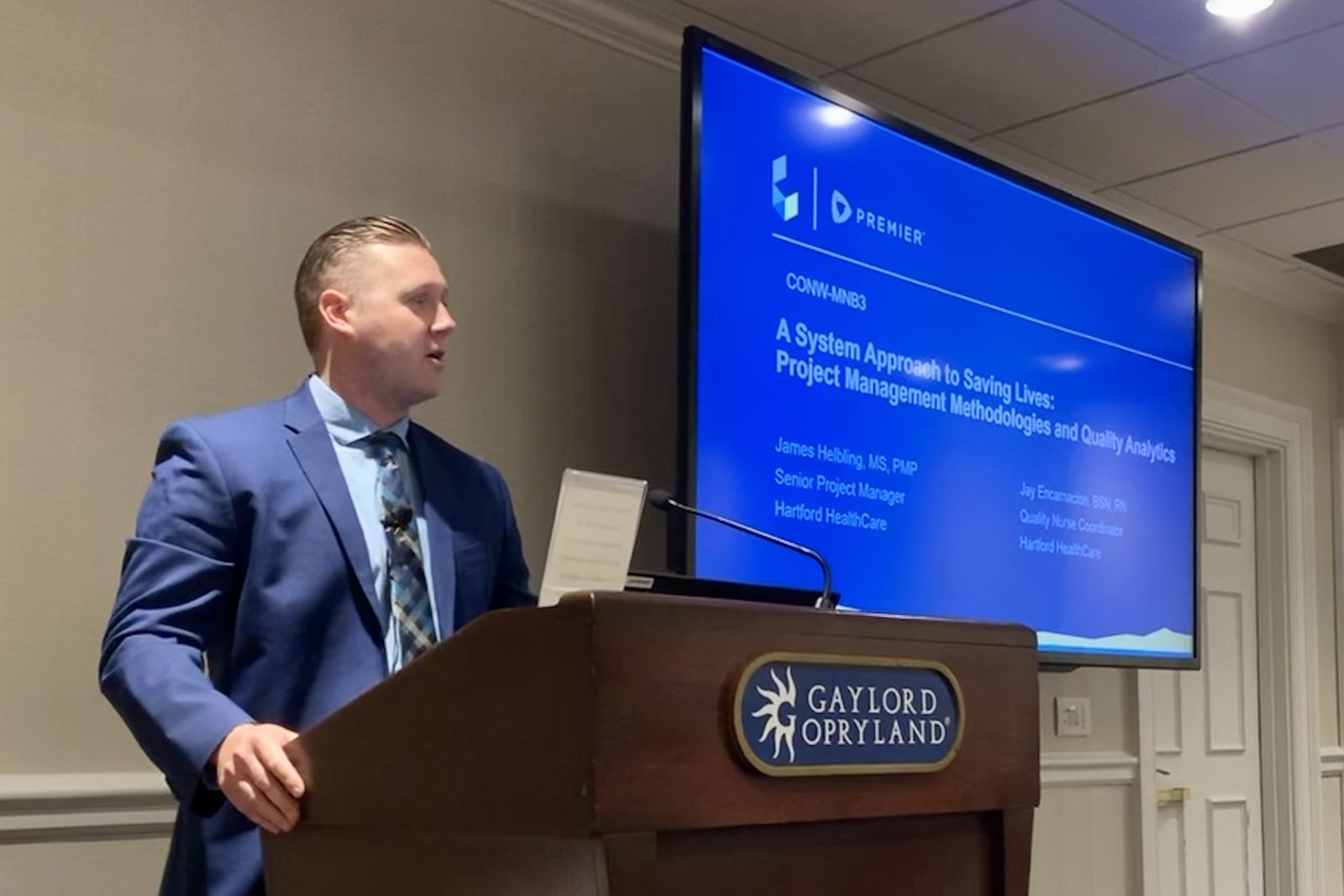 James Helbling speaking at a podium beside a presentation slide titled A System Approach to Saving Lives.